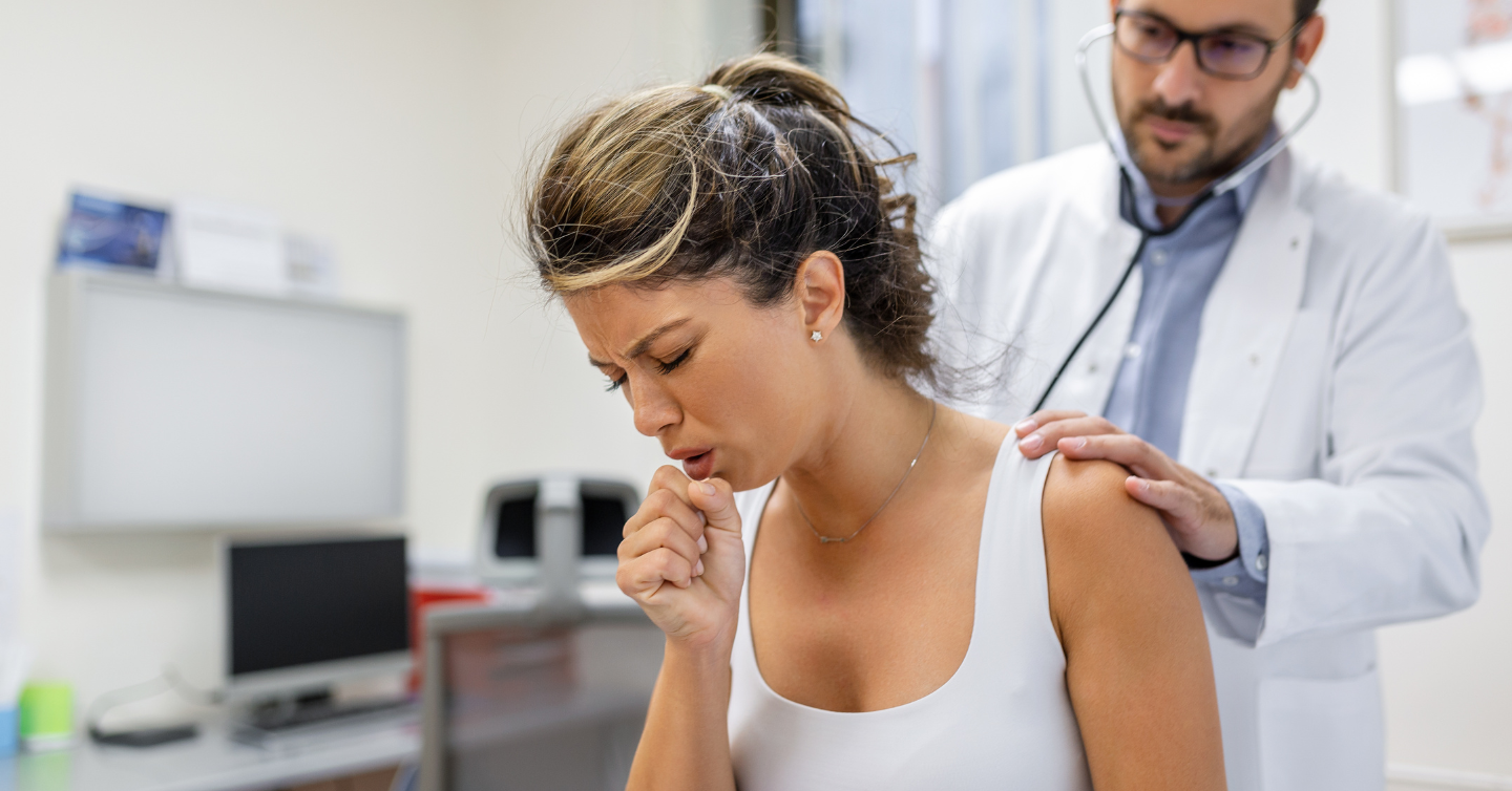 Mulher jovem de regata branca e tossindo enquanto um médico a examina com um estetoscópio