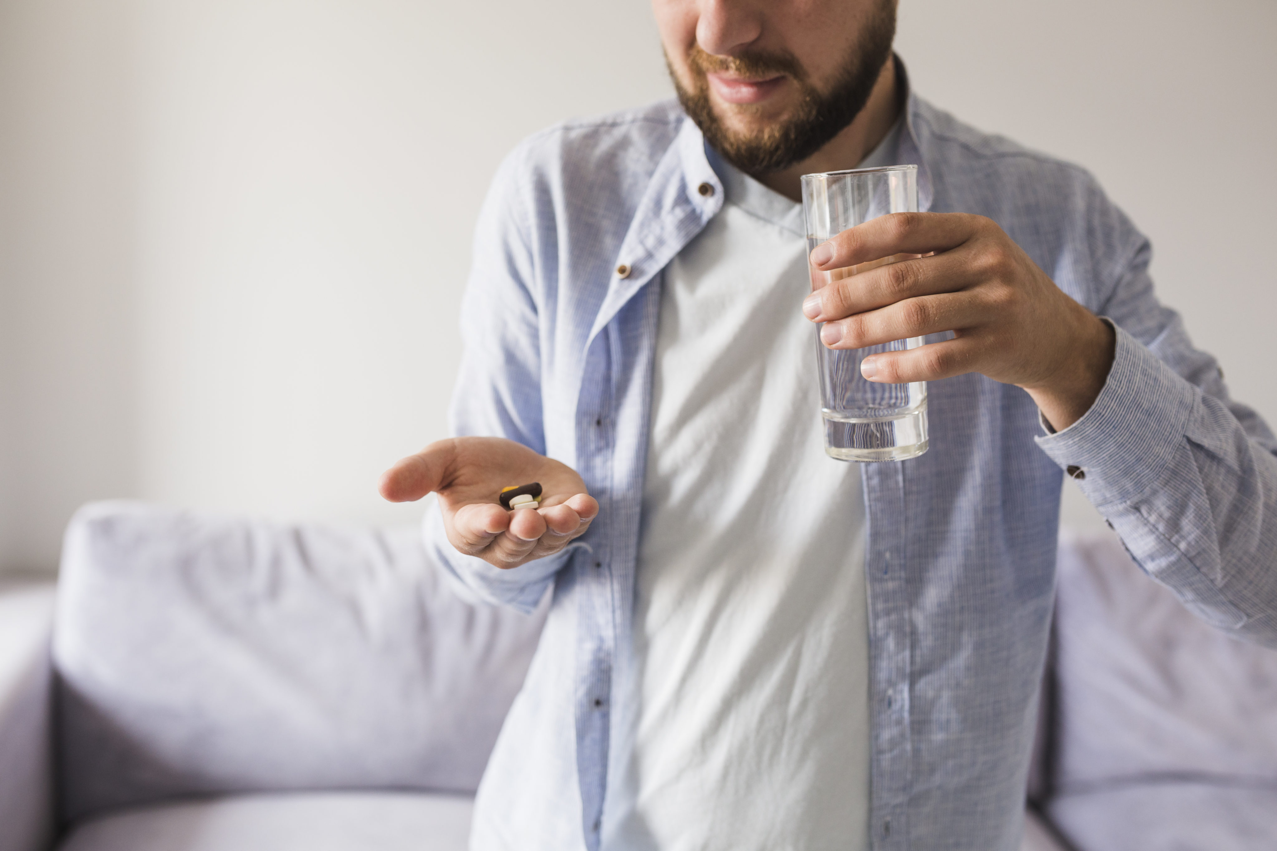 homem de pele branca, veste camiseta branca e camisa azul aberta por cima. Na mão direita segura alguns medicamentos e na mão esquerda um copo de vidro com água.