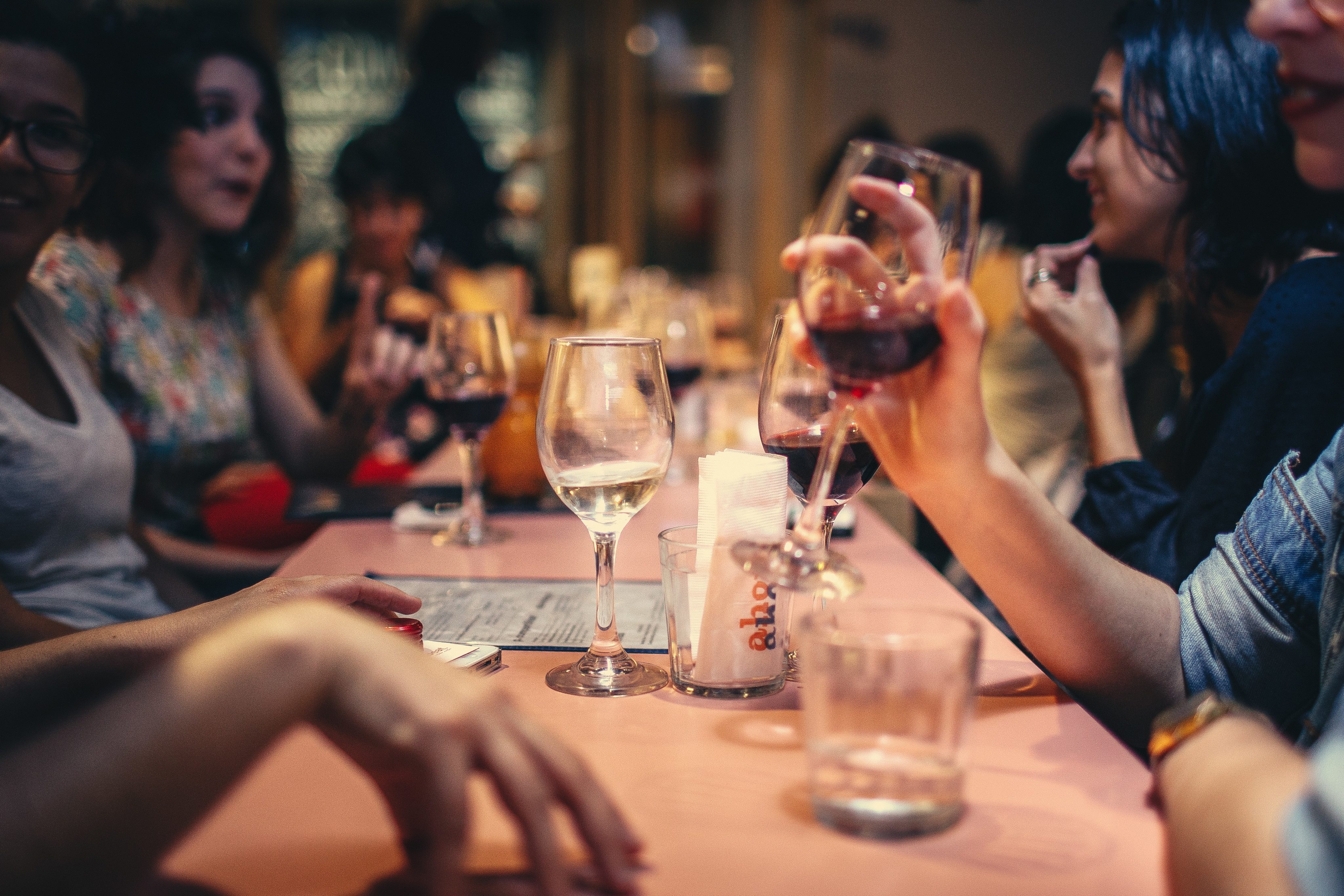 pessoas diversas sentadas em fundo. imagem foco tem duas pessoas, sem identificar gênero, sentados a mesa. As pessoas seguram, cada uma, uma taça de vidro com vinho.