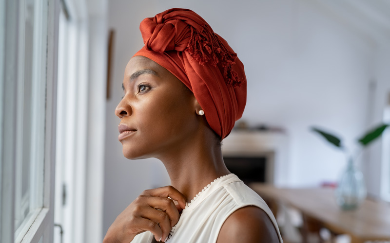 Mulher negra com um lenço na cabeça olhando pela janela de uma sala de estar